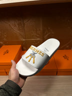 White Hermes slide sandal held up, gold logo on upper strap