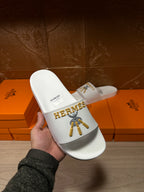 White Hermes slide sandal in hand, gold Hermes logo on front strap