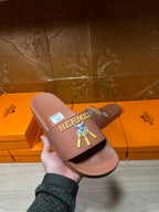 Hermes taupe slide sandal held in hand, multicolor logo patch on strap