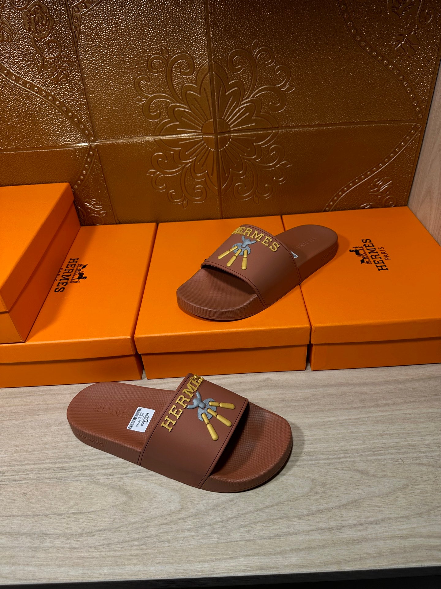 Hermes taupe slide sandals on floor, multicolor logo patch on single upper