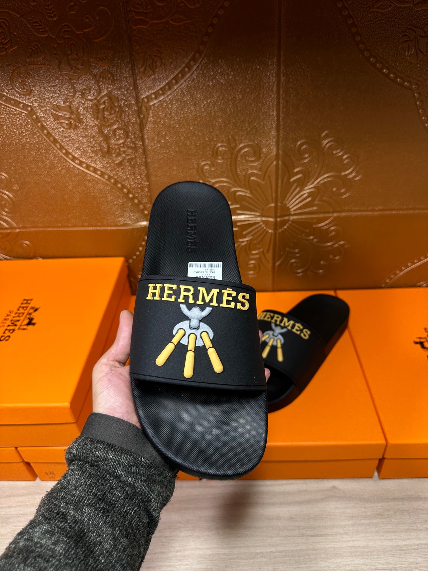 Hand holding a Hermes black slide sandal, showing front logo and white size label.