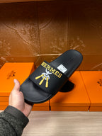 Hand holding Hermes black slide sandals with gold-and-blue logo on the strap.