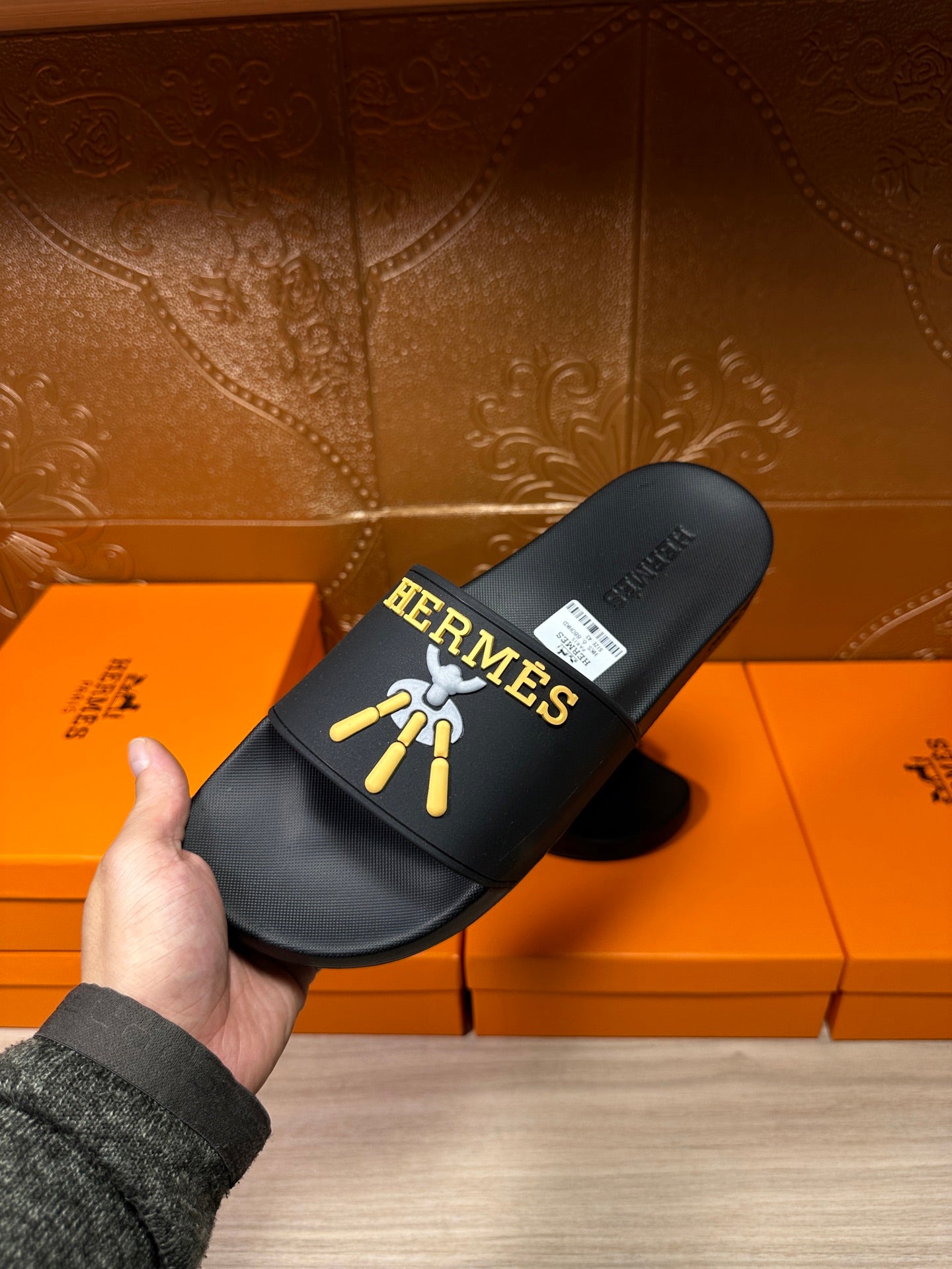 Hand holding Hermes black slide sandals with gold-and-blue logo on the strap.