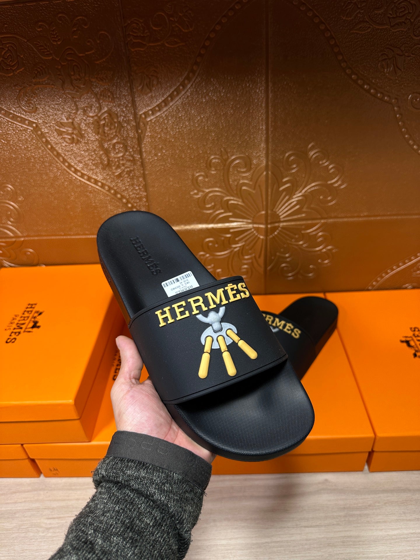 Hand holding Hermes black slide sandals with logo graphic on the front strap.