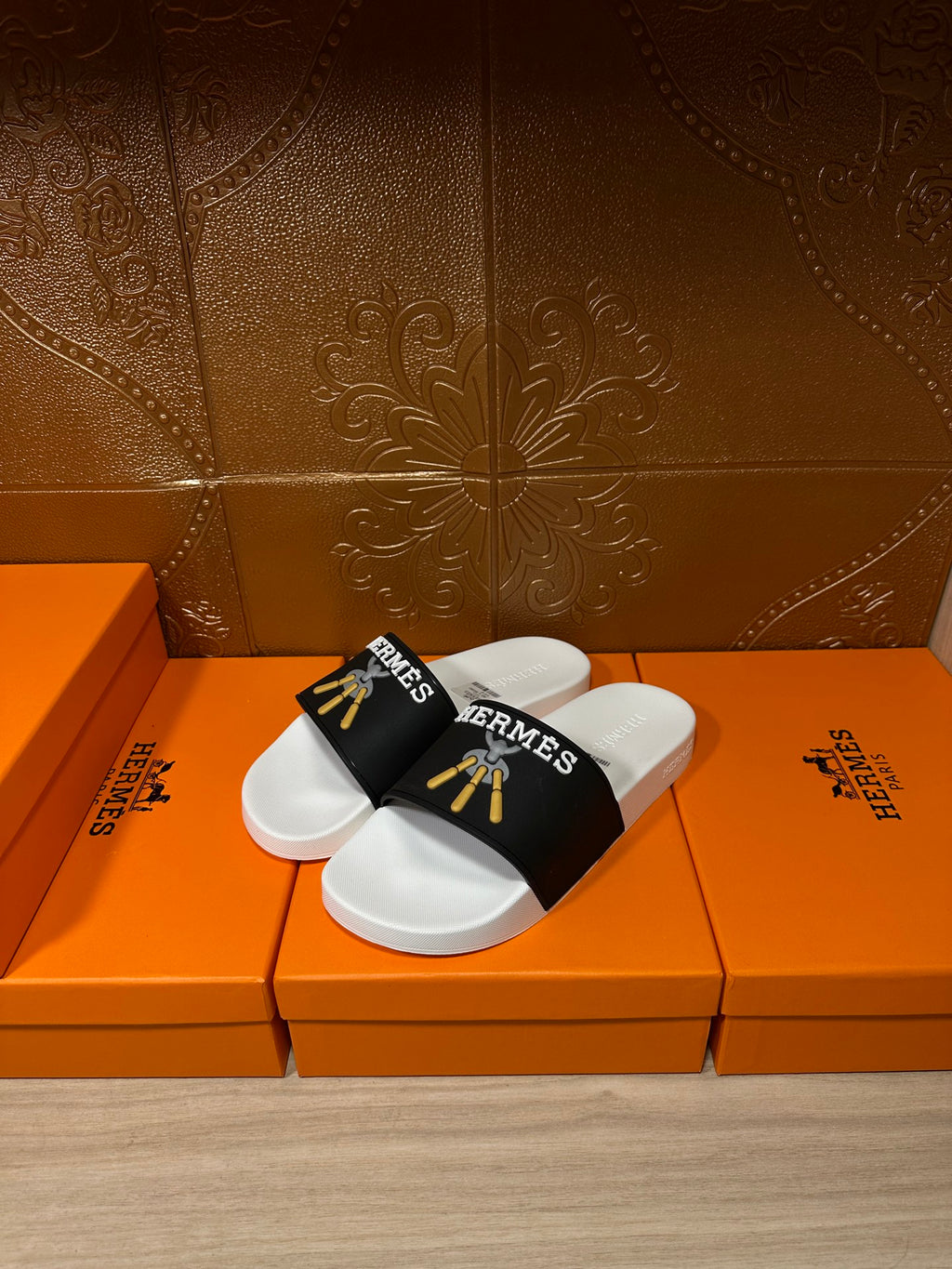 White slide sandals with black strap featuring Hermes logo and gold tassel detail