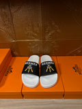 White slide sandals with black Hermes strap and gold tassel detail