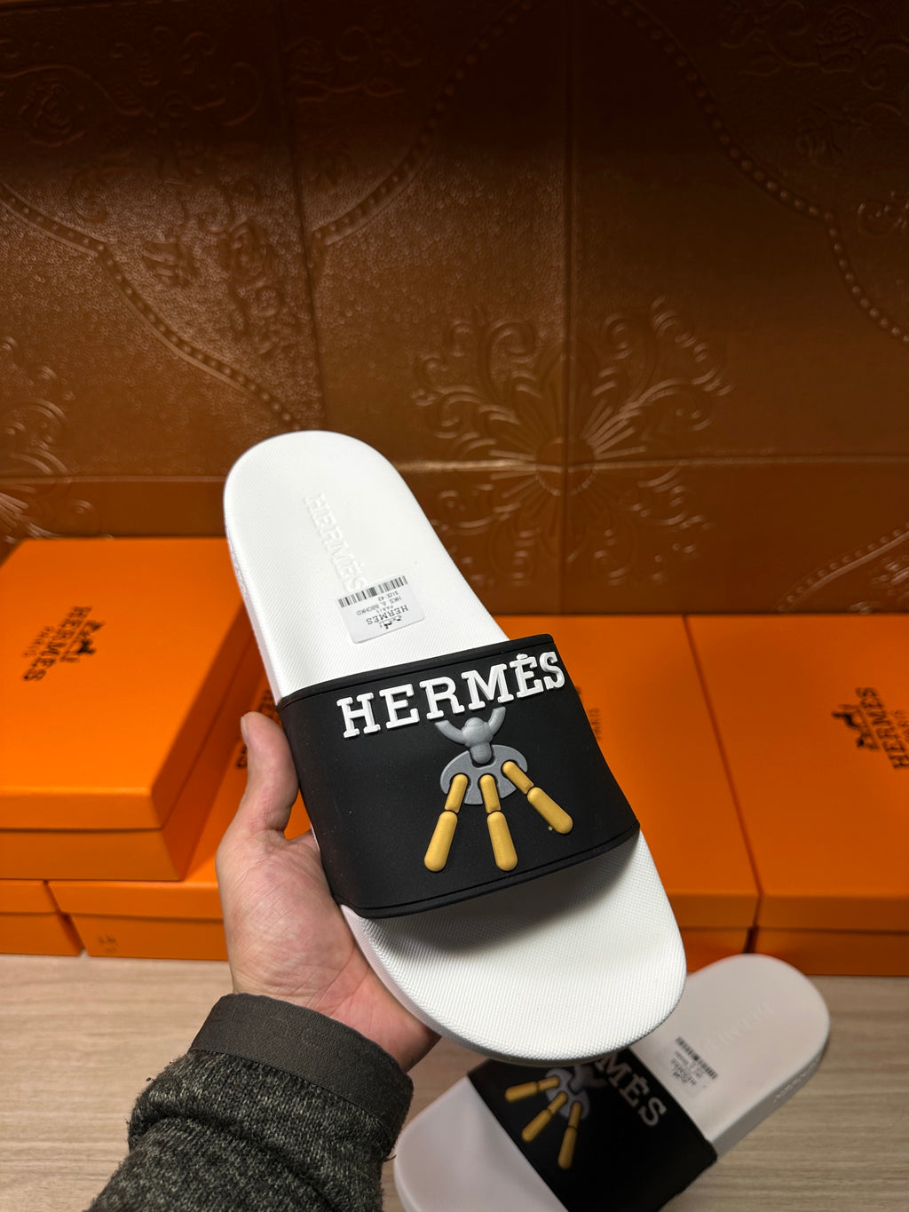 Handheld white slide sandal with black Hermes strap and gold tassel graphic