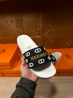 Hermès slide sandal held in hand: black logo strap on white footbed.