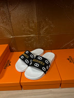 Hermès sandals on white footbeds with black strap featuring repeating logo.