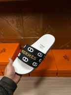 Hermès slide sandal held in hand, white sole with black strap and logo print.