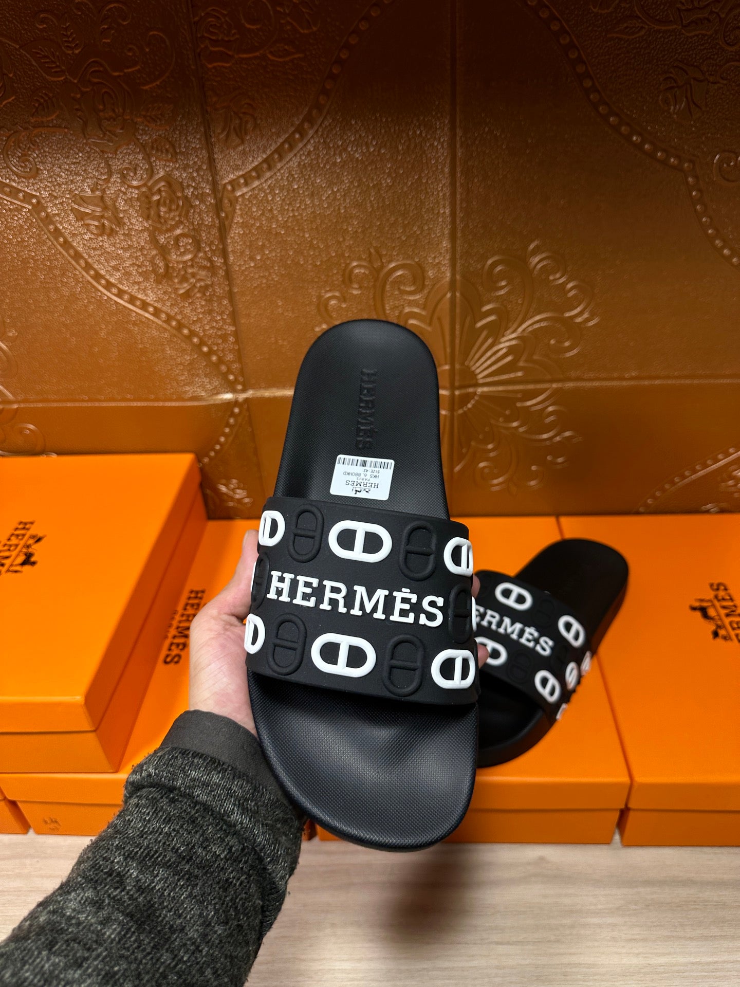 Black slide sandals held in hand, HERMES logo strap with white “H” lettering