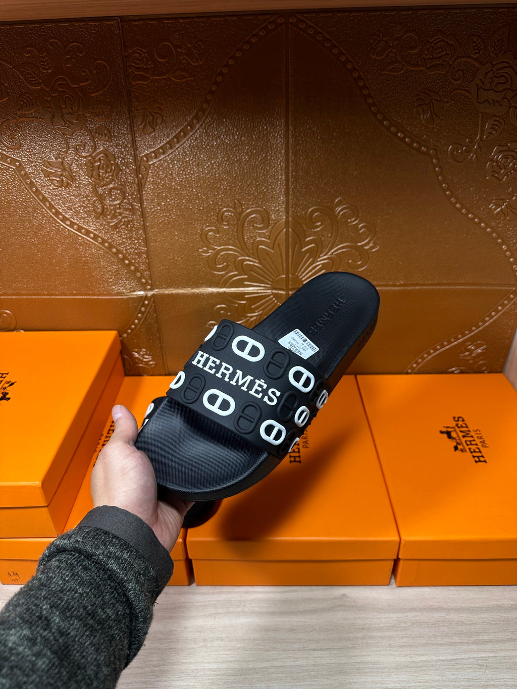Black slide sandals held in hand, wide HERMES logo strap on black footbed