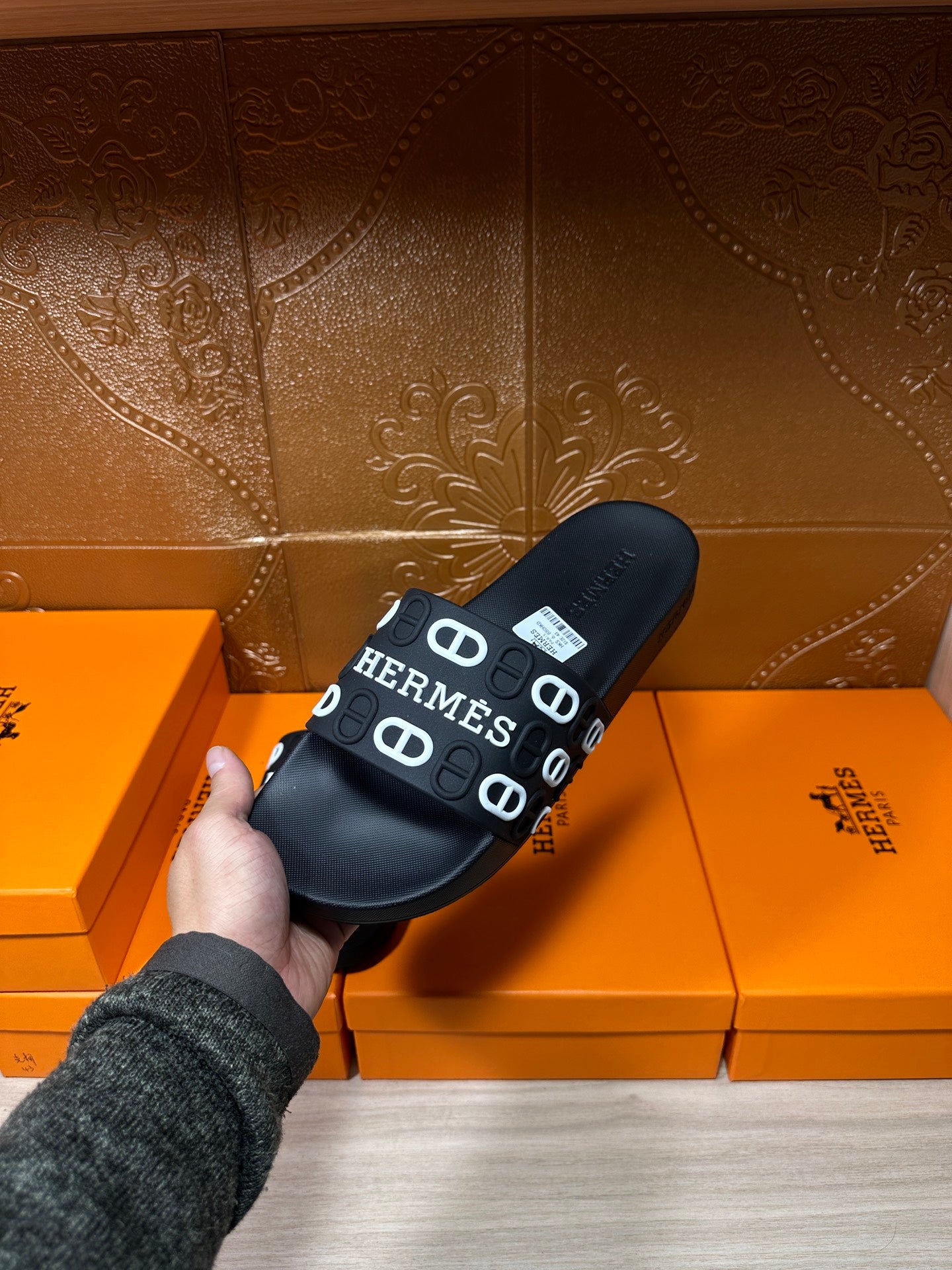 Black slide sandals held in hand, wide HERMES logo strap on black footbed