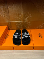 Black slide sandals with wide logo strap, white “HERMES” print and buckle details