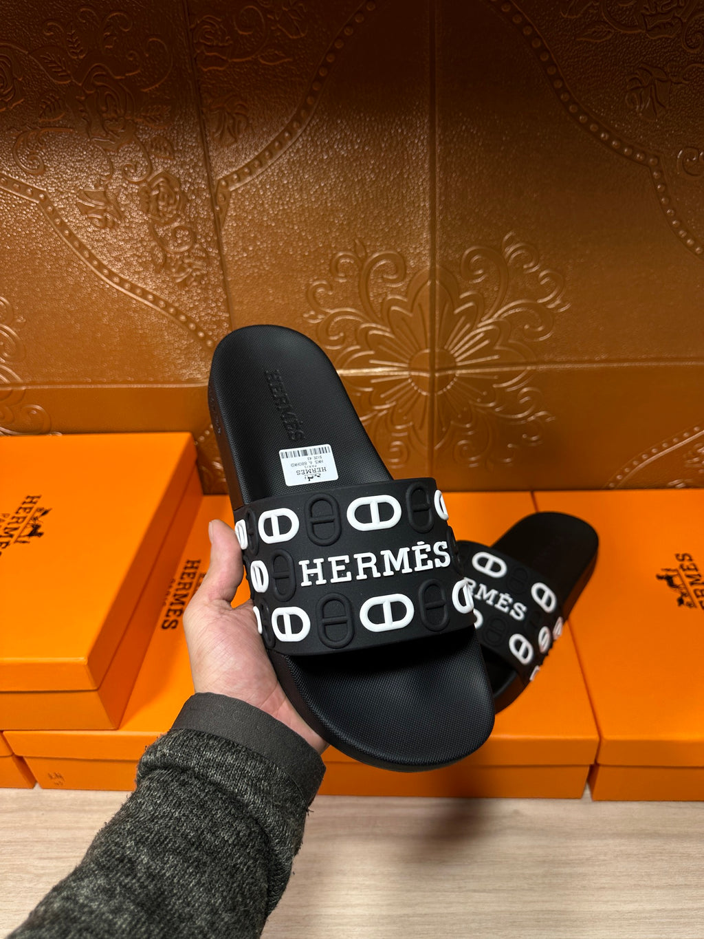 Black slide sandals held in hand, wide HERMES logo strap with white motif loops