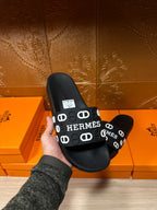 Black slide sandals held in hand, wide HERMES logo strap with white motif loops