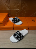White slide sandal with black strap showing Hermes logos, side view