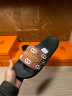 Black slide sandal held in hand, brown Hermes logo strap on top