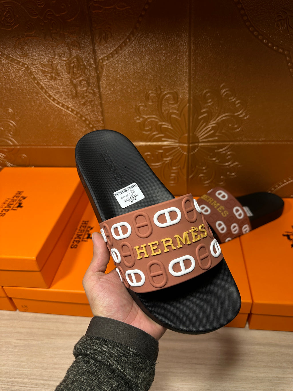Black slide sandal held up, brown Hermes logo strap and label on footbed