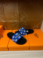 Black slide sandals with blue Hermes-pattern strap and open-toe silhouette.
