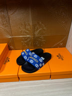 Black slide sandals with blue Hermes-logo patterned strap, front angled view.