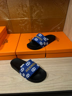 Black slide sandals with blue Hermes-pattern strap, pair shown on the floor.