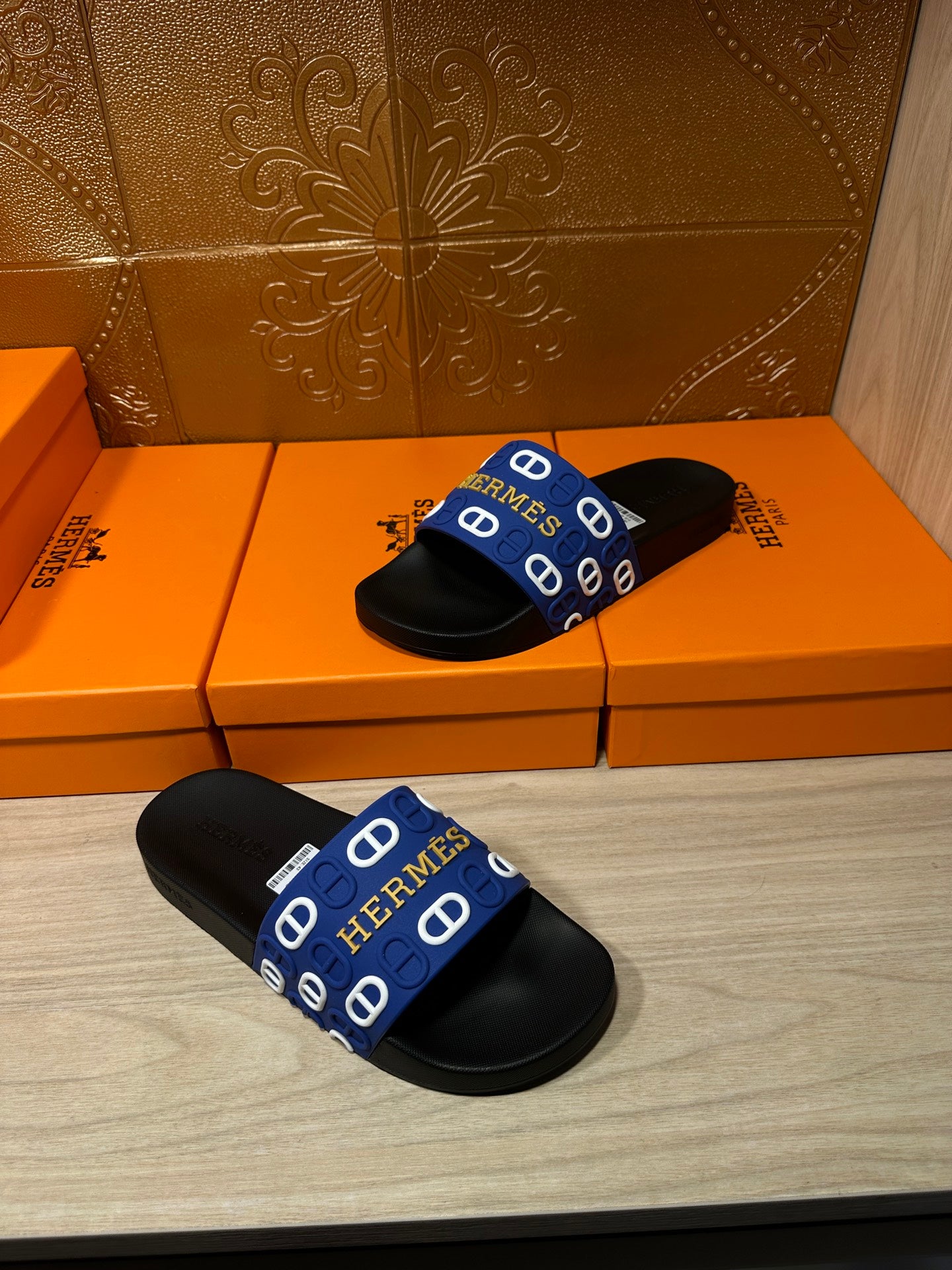 Black slide sandals with blue Hermes-pattern strap, pair shown on the floor.
