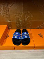 Black slide sandals with blue Hermes-pattern strap, flat footbed and wide band.