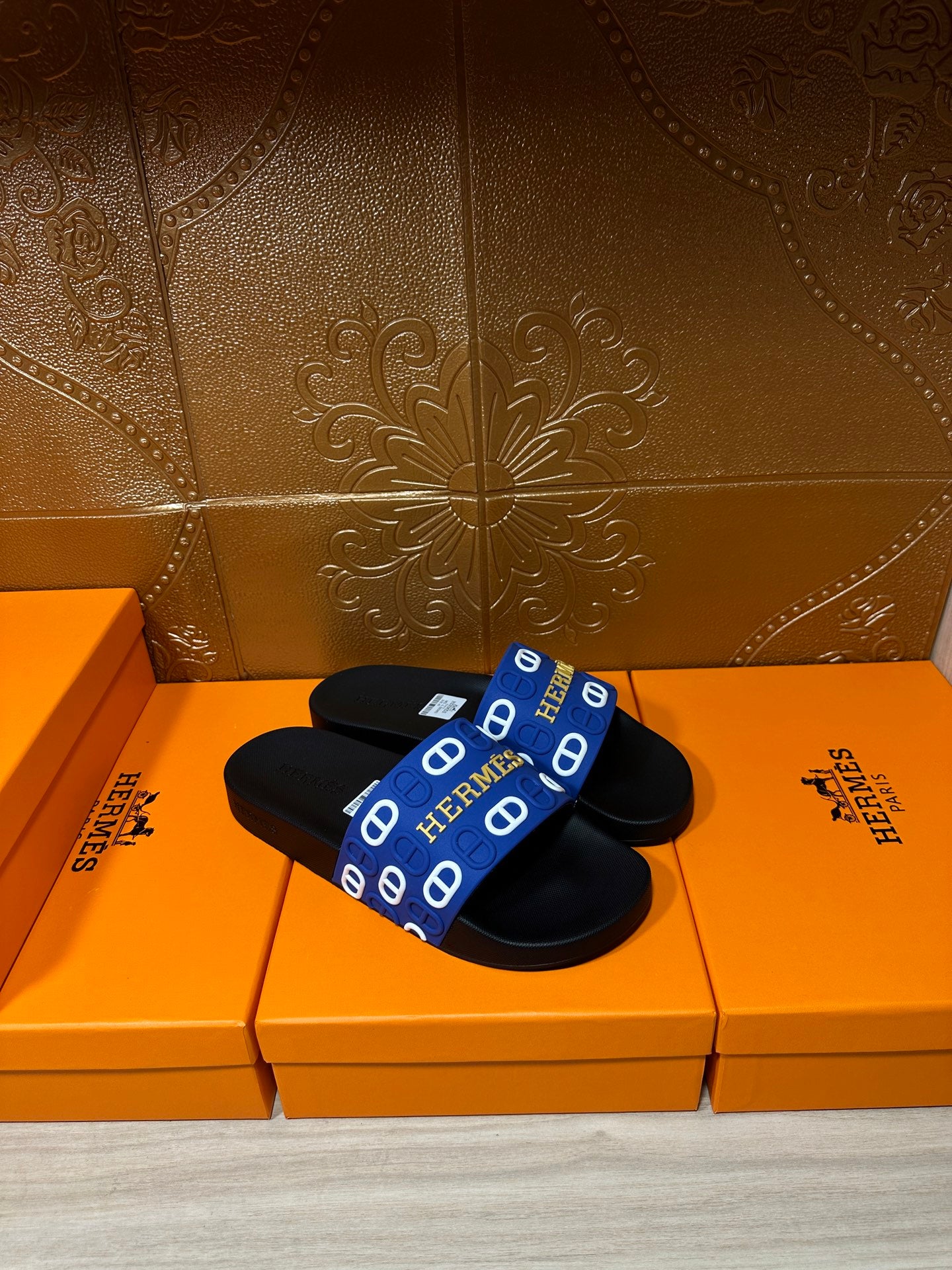 Black slide sandals with blue wide strap featuring repeated Hermes logo motifs.