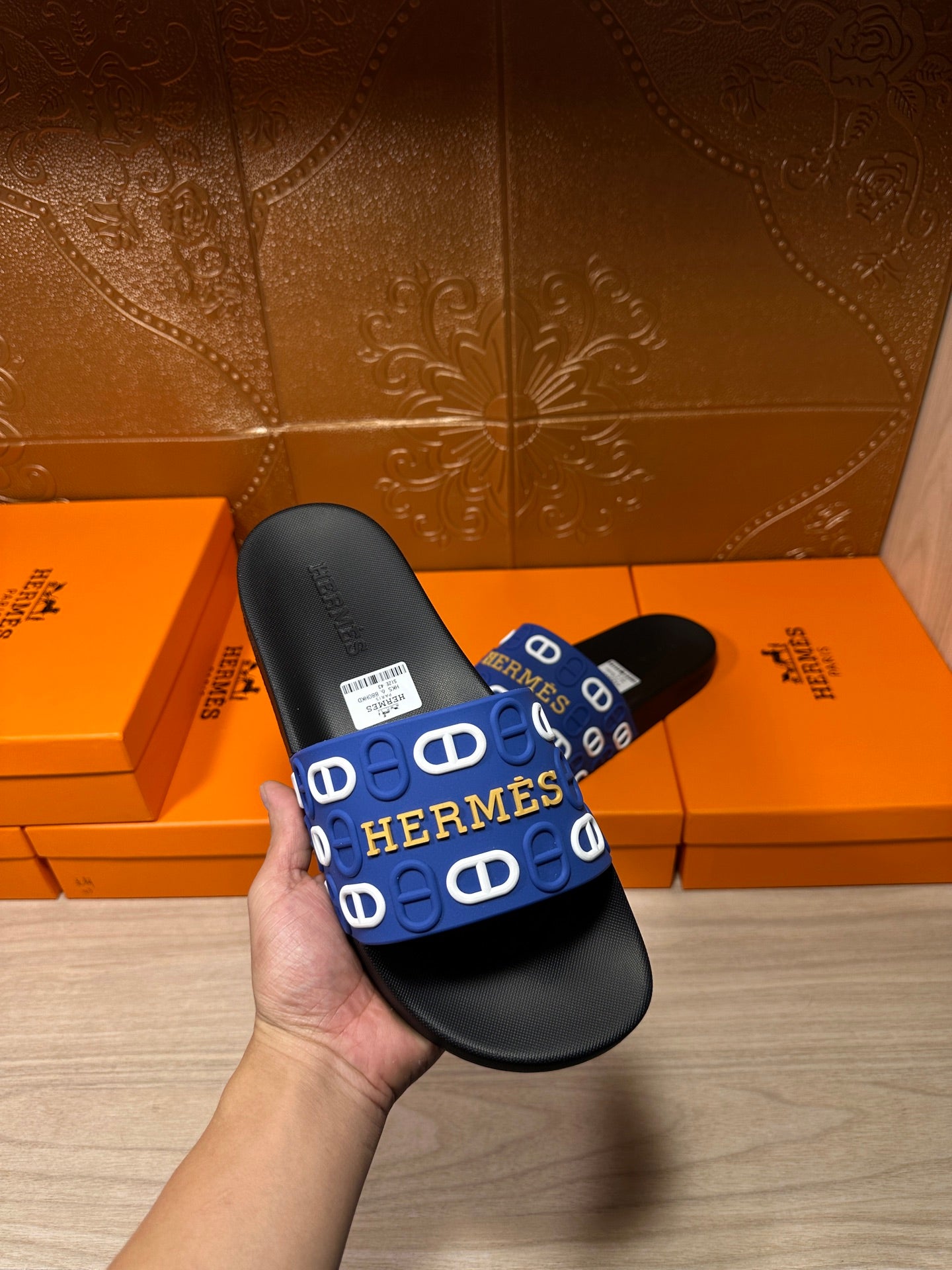 Black slide sandal held in hand, blue wide strap with repeated Hermes logo motifs.