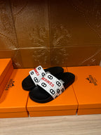 Black slide sandals with white patterned strap and Hermes logo.