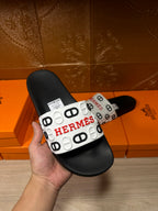 Black slide sandal with white Hermes logo strap, held in a hand.
