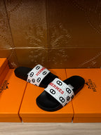 Black slide sandals with white logo band across the foot, Hermes.