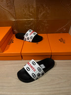 Black slide sandals with white Hermes logo strap, laid on display boxes.