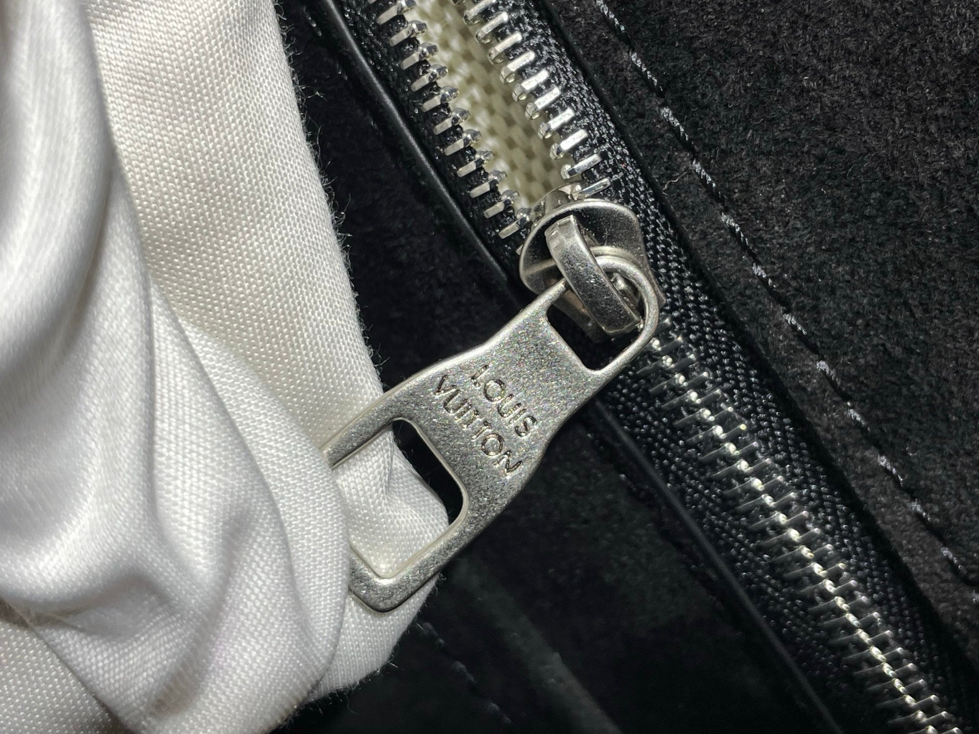 Metal zipper pull with logo tag on black bag, white cloth partly visible