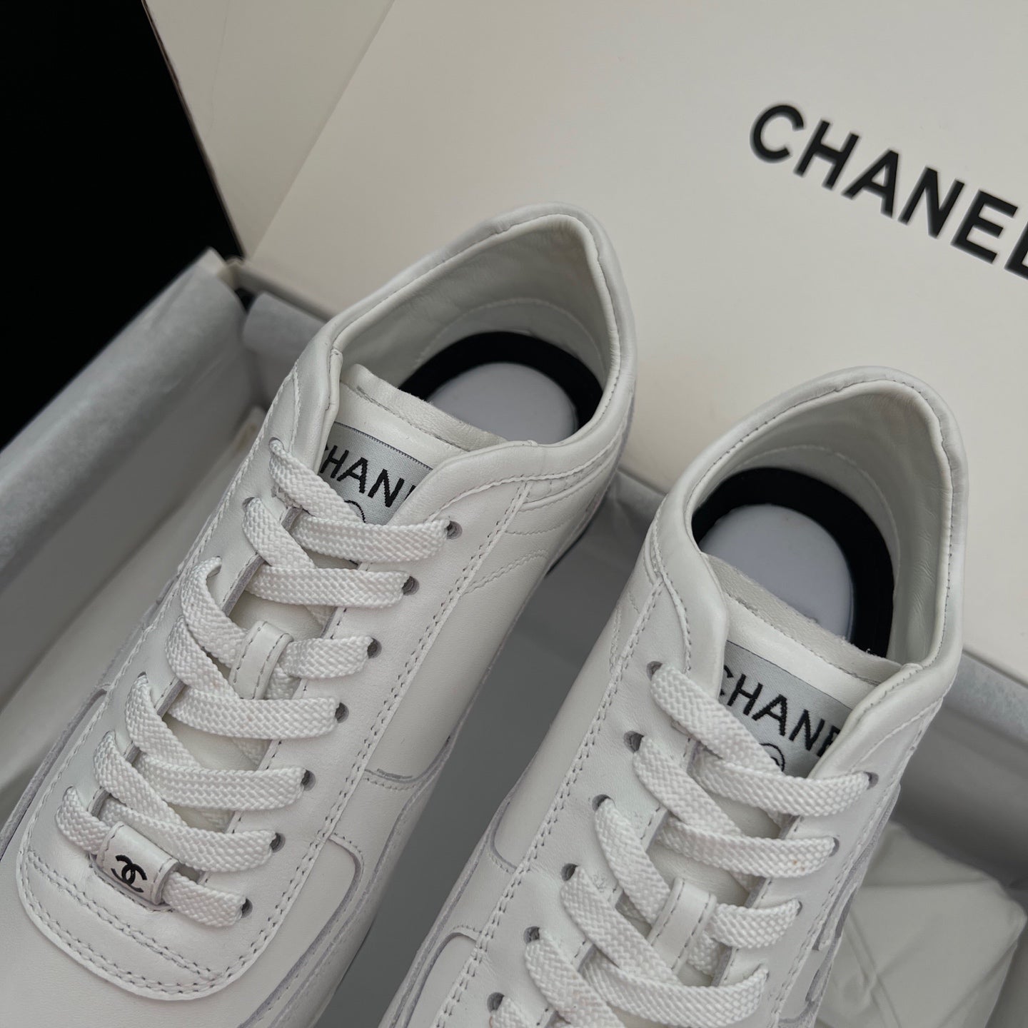 Close-up of white Chanel unisex lace-up sneakers with stitched logo detail.
