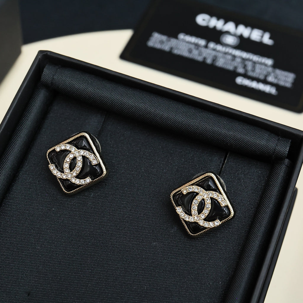 Pair of square logo stud earrings with black enamel and crystal details on dark display tray