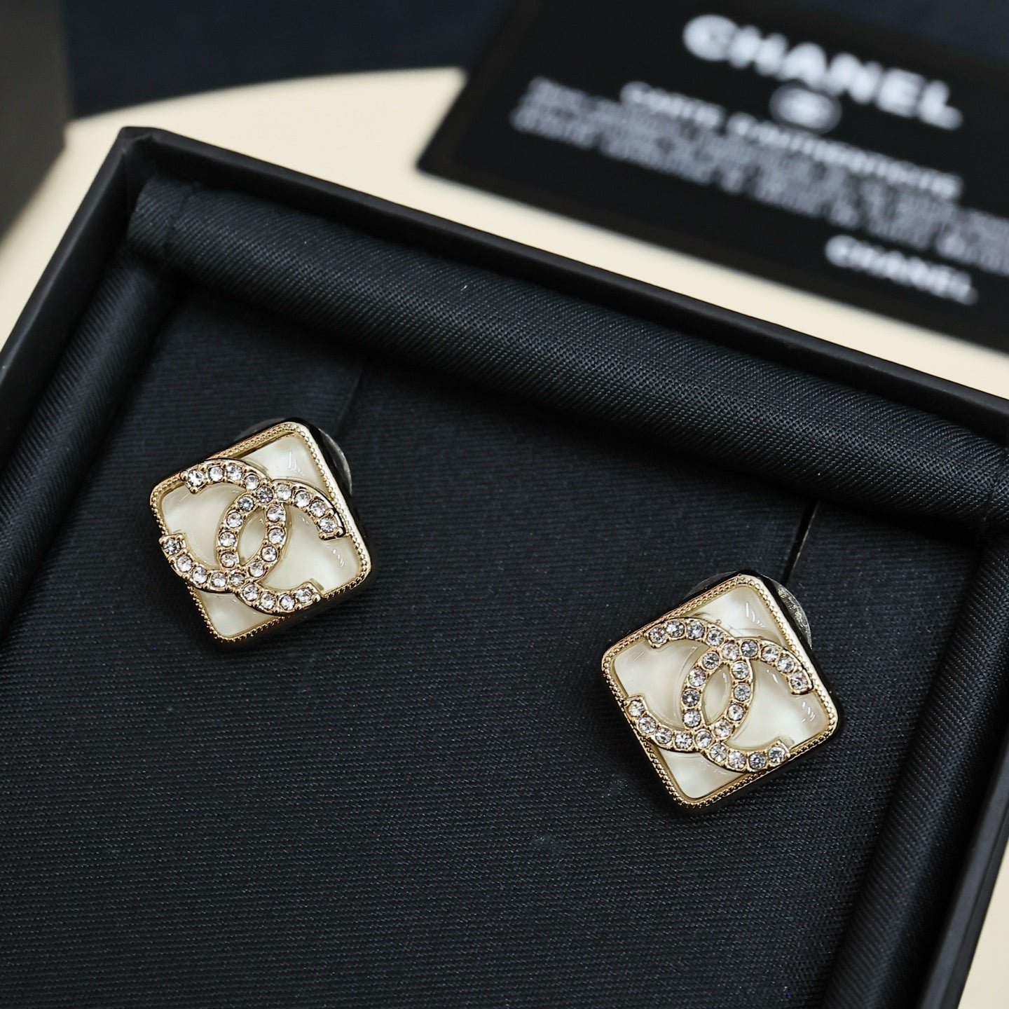 Square stud earrings with white logo panel, gold-tone border, and sparkling crystal details