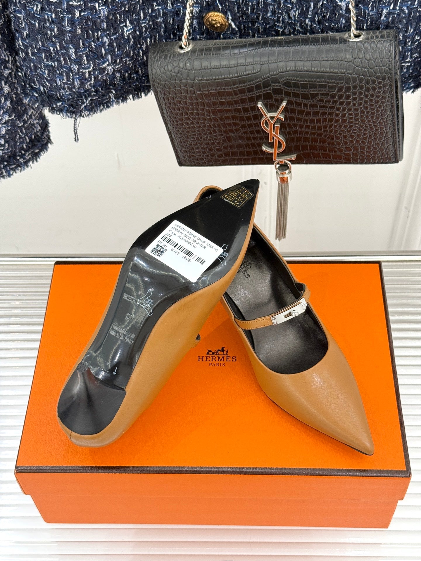 Tan pointed-toe heeled pumps with ankle strap, product tag on shoe, orange box.
