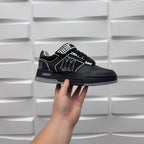 Black low-top sneaker with checkerboard strap detail and graphic logo, held by hand.