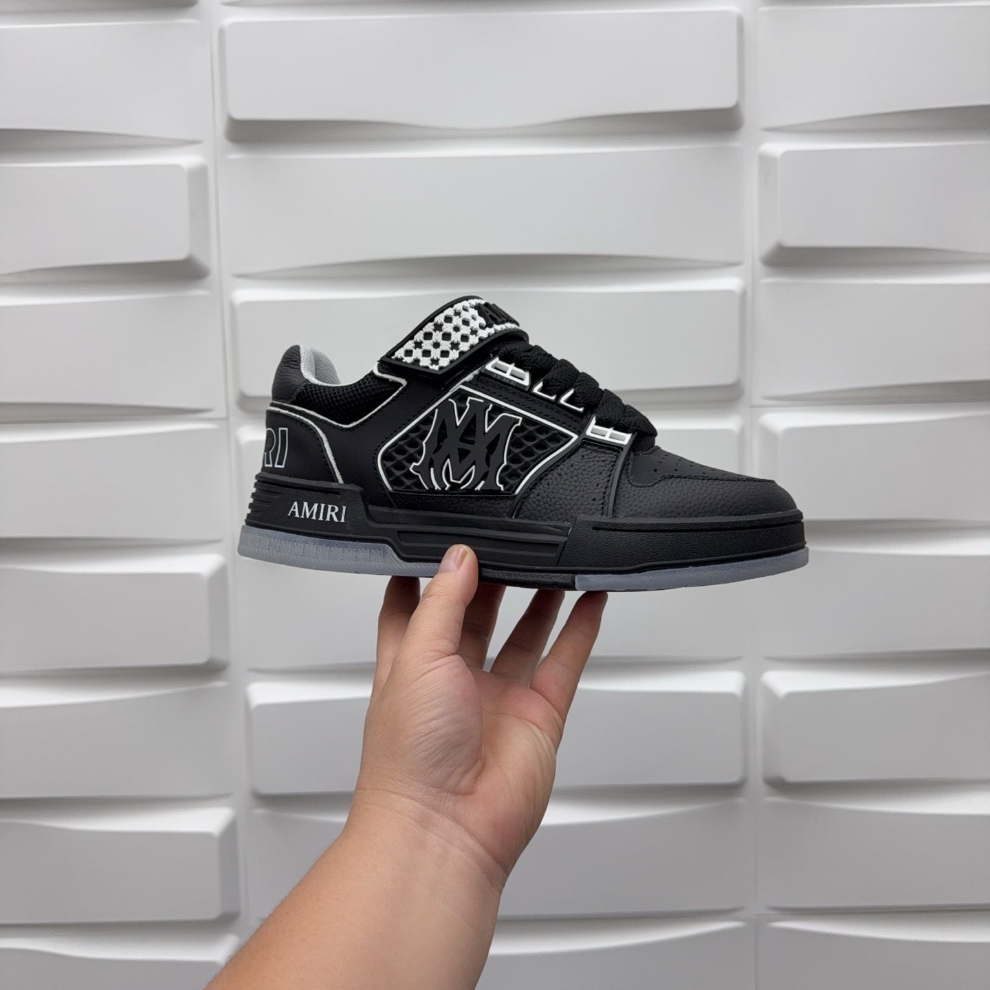 Black low-top sneaker with checkerboard strap detail and graphic logo, held by hand.