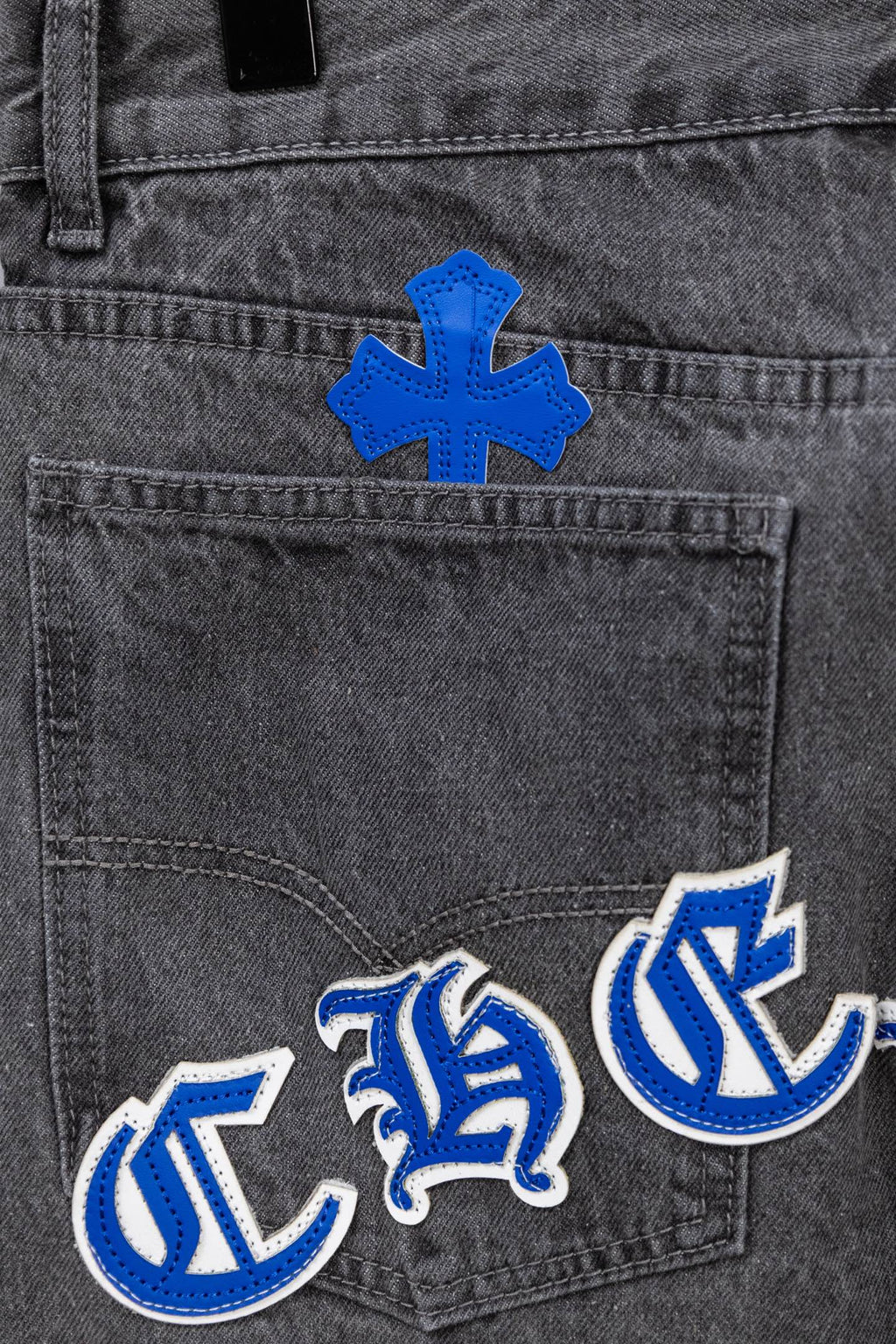 Gray denim pants close-up with blue cross patch and blue-and-white logo embroidery.