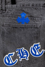 Gray denim pants close-up with blue cross patch and blue-and-white logo embroidery.
