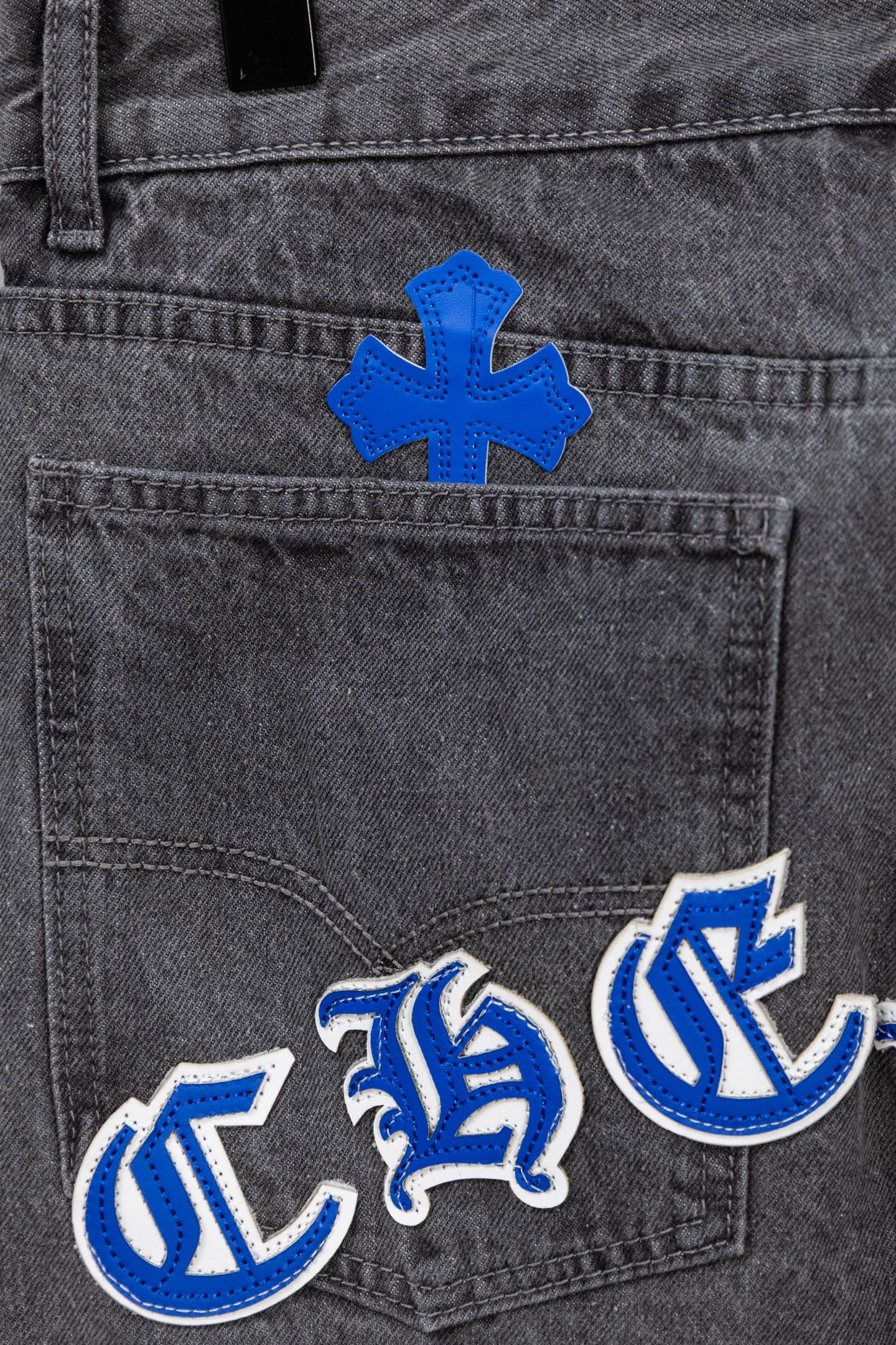 Gray denim pants close-up with blue cross patch and blue-and-white logo embroidery.
