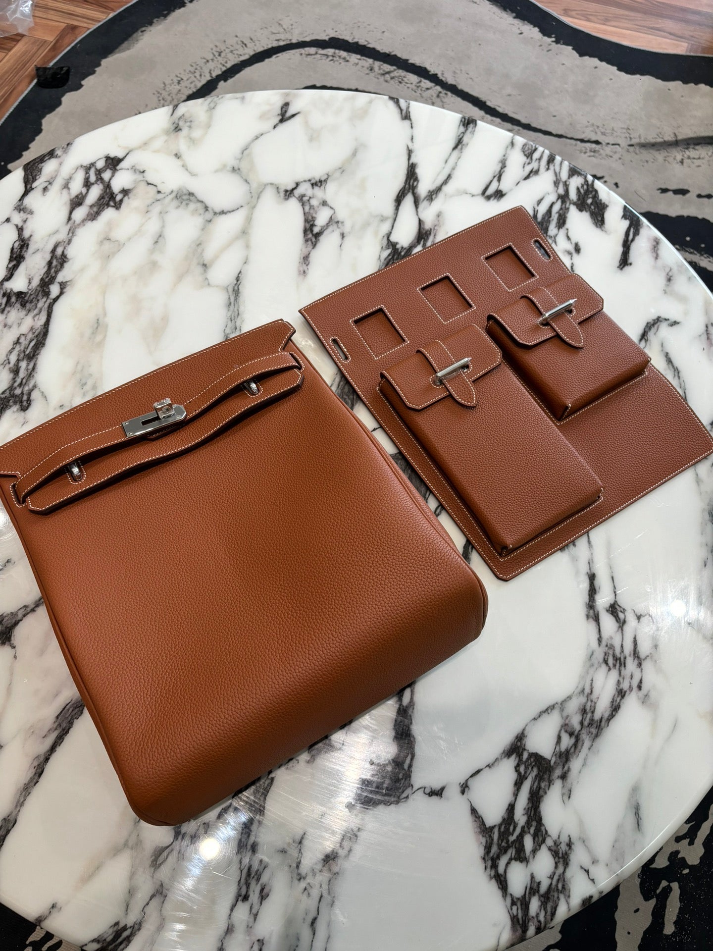 Pair of Hermes bags in tan brown leather with front flap, silver buckles, and stitched edges.