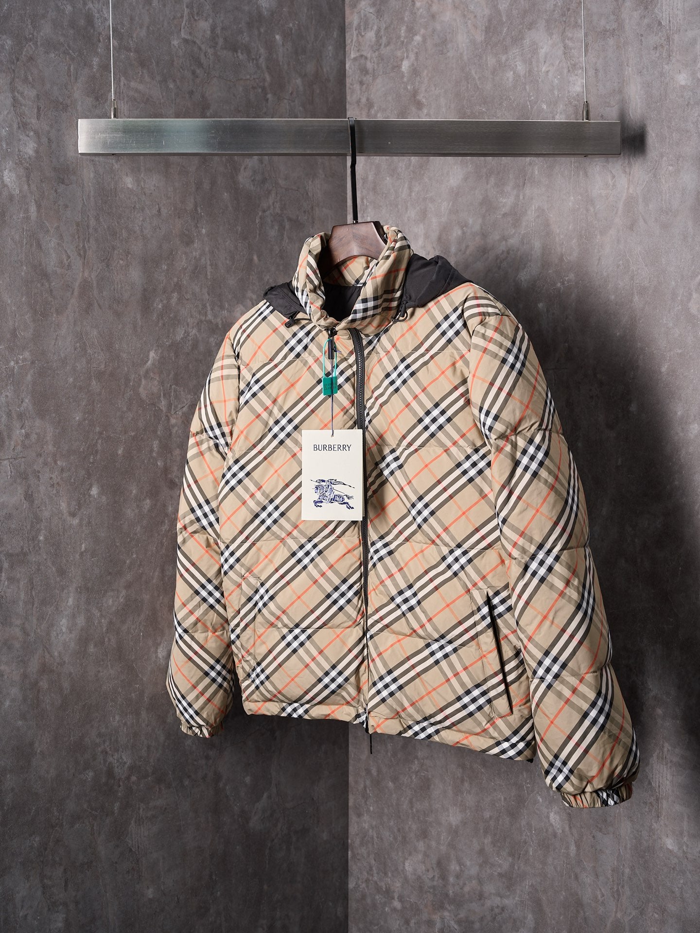 Plaid puffer jacket with black hood and front zip, featuring a hang tag at chest.