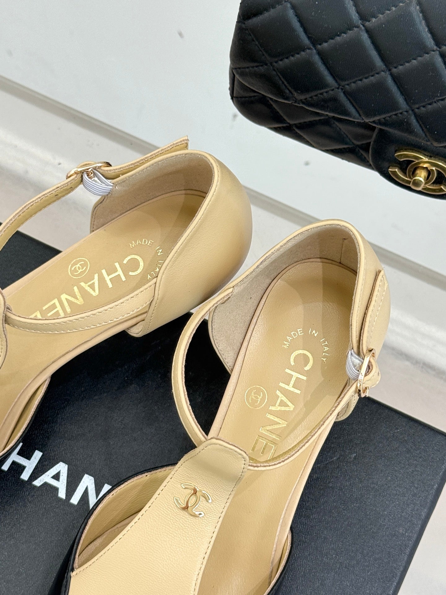 Beige pointed-toe slingback heels with ankle strap and logo detailing, on box.