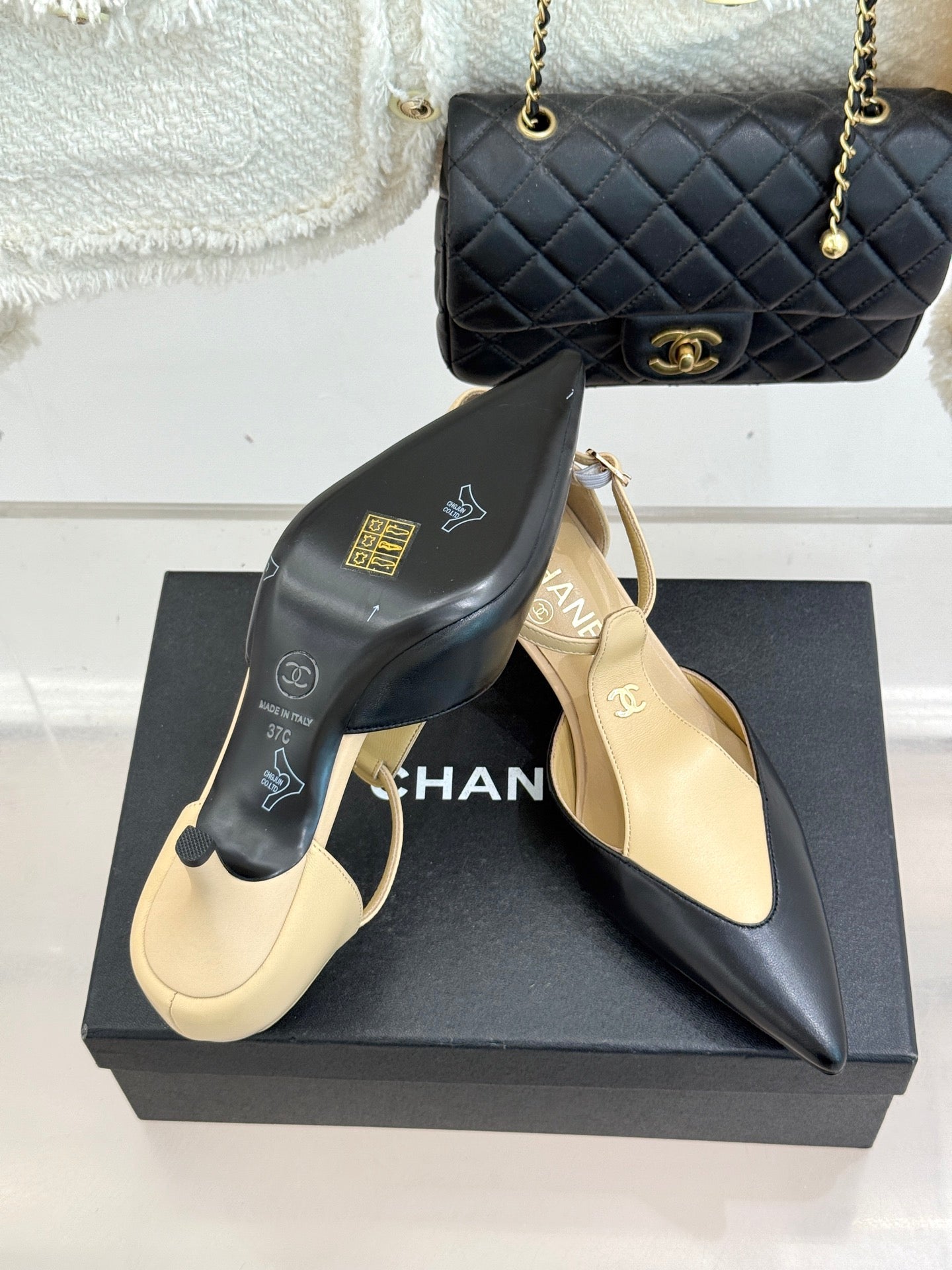 Beige and black pointed-toe slingback heels with ankle straps, on display box.