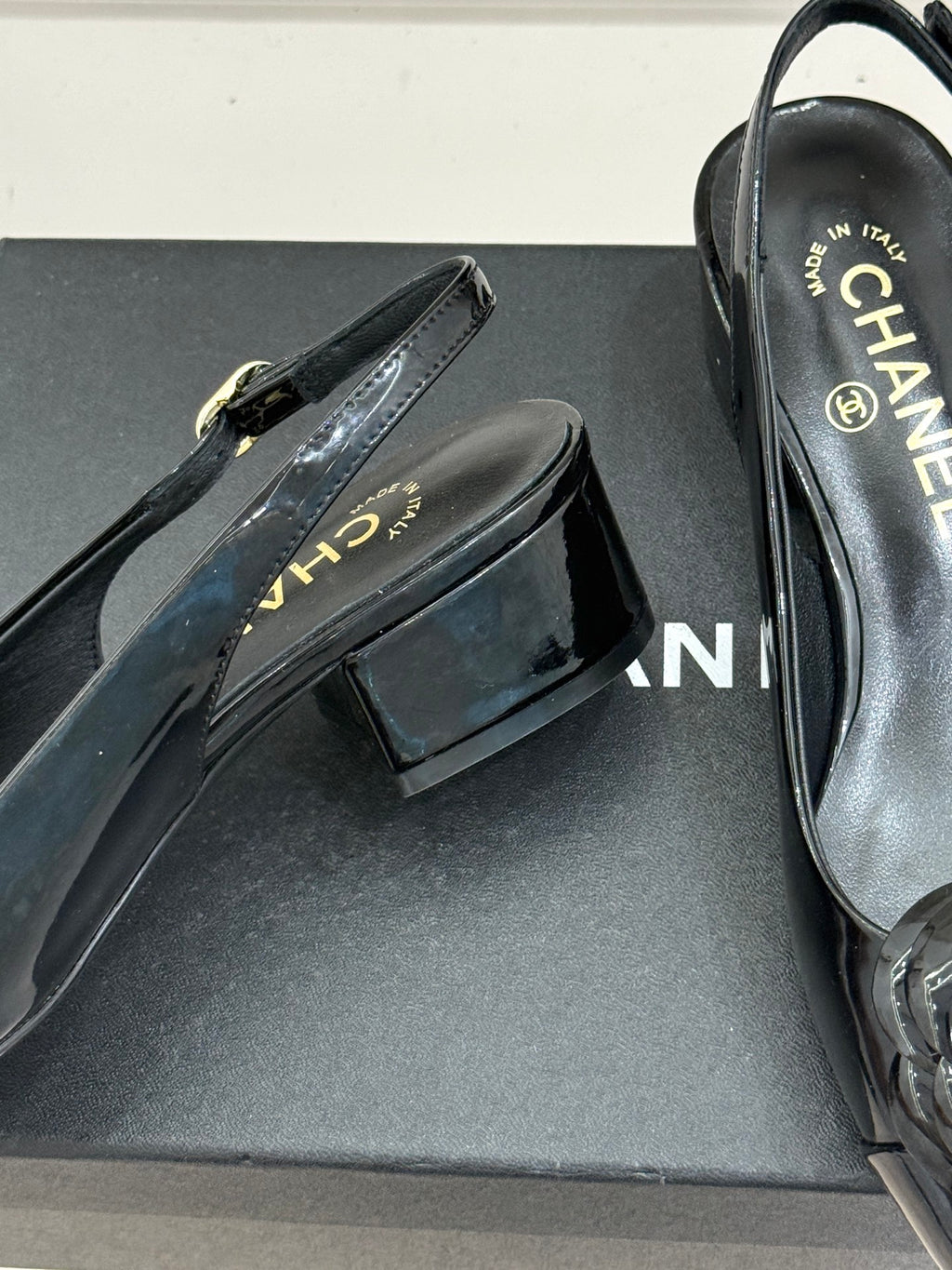 Black patent leather flat shoe with gold-tone buckle strap on a dark shoe box