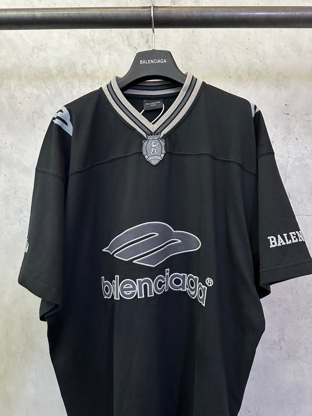 Black short-sleeve shirt with V-neck trim, crest charm at collar, and logo graphic